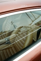 A close-up view through the side window of a car, showcasing a brown leather seat with visible reflections on the glass. The seat has a retro design with horizontal stitching, and the exterior of the car appears to be a muted red or maroon color.