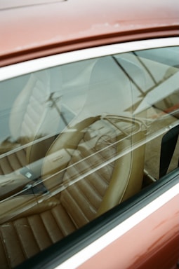 A close-up view through the side window of a car, showcasing a brown leather seat with visible reflections on the glass. The seat has a retro design with horizontal stitching, and the exterior of the car appears to be a muted red or maroon color.