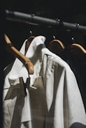 A close-up view of several light-colored shirts on wooden hangers. The hangers are arranged on a metal rod against a dark background, with soft lighting creating a calm and minimalistic atmosphere.