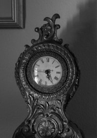 An intricately carved antique clock with Roman numerals on its clock face. The clock has ornate details including floral patterns and scrollwork, made from dark, possibly wooden or metal material. The image is in black and white, highlighting the textures and craftsmanship of the clock against a plain wall.