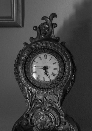 An intricately carved antique clock with Roman numerals on its clock face. The clock has ornate details including floral patterns and scrollwork, made from dark, possibly wooden or metal material. The image is in black and white, highlighting the textures and craftsmanship of the clock against a plain wall.