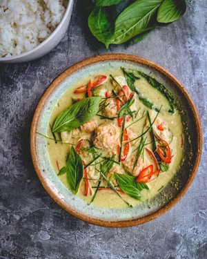 A bowl of green curry with shrimp, garnished with fresh basil leaves, sliced red chili peppers, and finely cut herbs. The dish is placed on a textured gray surface next to a bowl of white rice and additional basil leaves.