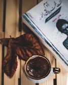 A cup of coffee rests on a wooden surface beside a book with Arabic text on the cover. Two dried leaves lie next to the cup and book, creating an autumnal feel.