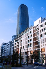 A modern cityscape featuring a tall, cylindrical glass skyscraper towering over an adjacent multi-story building with a mix of glass and brick elements. Several flags are displayed in front of the building, and trees line the street. Cars are parked along the road, and a few pedestrians are visible.