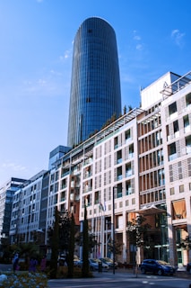 A modern cityscape featuring a tall, cylindrical glass skyscraper towering over an adjacent multi-story building with a mix of glass and brick elements. Several flags are displayed in front of the building, and trees line the street. Cars are parked along the road, and a few pedestrians are visible.