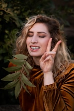 A person with curly blonde hair is smiling with closed eyes, holding a peace sign with one hand and a green leaf near their face with the other. They are wearing a brown outfit and standing against a backdrop of lush green foliage.
