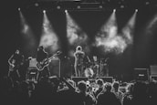 A black-and-white photograph of a live concert performance with four musicians on stage. The central figure is a singer holding a microphone stand with both hands. To the left, a guitarist and a bassist are playing their instruments. On the right, a drummer is positioned behind a drum set. There's a crowd of people in the foreground, some capturing the event with their phones. Stage lights illuminate the scene from above, casting beams of light and creating a smoky atmosphere.