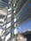 An architectural scene featuring modern structures with large glass windows and horizontal slats casting shadows. The scene includes reflections of the sky and urban buildings in the background with graffiti visible on some of the exterior surfaces.
