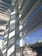 An architectural scene featuring modern structures with large glass windows and horizontal slats casting shadows. The scene includes reflections of the sky and urban buildings in the background with graffiti visible on some of the exterior surfaces.