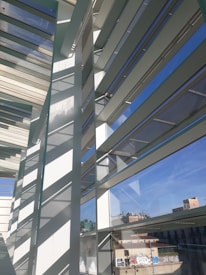 An architectural scene featuring modern structures with large glass windows and horizontal slats casting shadows. The scene includes reflections of the sky and urban buildings in the background with graffiti visible on some of the exterior surfaces.
