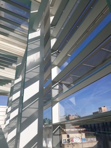 An architectural scene featuring modern structures with large glass windows and horizontal slats casting shadows. The scene includes reflections of the sky and urban buildings in the background with graffiti visible on some of the exterior surfaces.