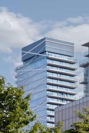 A modern high-rise building with a sleek glass facade and geometric design elements. The structure features multiple balconies and reflective windows, giving it a contemporary urban feel. The sky is bright with fluffy white clouds, and there are green tree leaves partially framing the lower part of the image.