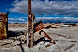 A desolate landscape with a rusty wooden structure and metal beams, partially collapsed and covered in graffiti. The ground is barren, and there are scattered pieces of debris. In the background, a range of mountains under a sky filled with textured clouds.