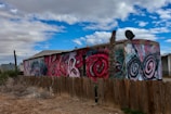 A mobile home with vibrant graffiti art featuring bold spirals and abstract designs on its side. The structure is set within a dusty landscape, enclosed by a wooden fence. A satellite dish is mounted on the roof, and a single bare tree rises alongside the building. The sky above is a mix of bright blue with scattered clouds.