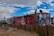 A mobile home with vibrant graffiti art featuring bold spirals and abstract designs on its side. The structure is set within a dusty landscape, enclosed by a wooden fence. A satellite dish is mounted on the roof, and a single bare tree rises alongside the building. The sky above is a mix of bright blue with scattered clouds.