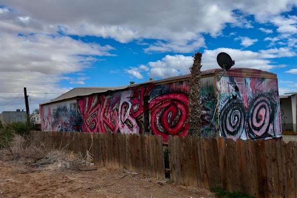 A mobile home with vibrant graffiti art featuring bold spirals and abstract designs on its side. The structure is set within a dusty landscape, enclosed by a wooden fence. A satellite dish is mounted on the roof, and a single bare tree rises alongside the building. The sky above is a mix of bright blue with scattered clouds.