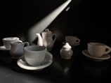 A collection of white porcelain cups, saucers, a teapot, and a milk jug are arranged on a dark surface. A beam of light from the top right casts dramatic shadows, highlighting the glossy surfaces of the ceramic pieces.