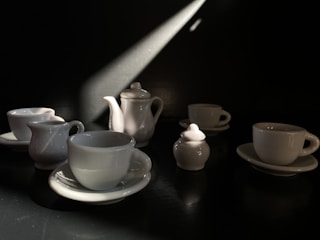 A collection of white porcelain cups, saucers, a teapot, and a milk jug are arranged on a dark surface. A beam of light from the top right casts dramatic shadows, highlighting the glossy surfaces of the ceramic pieces.