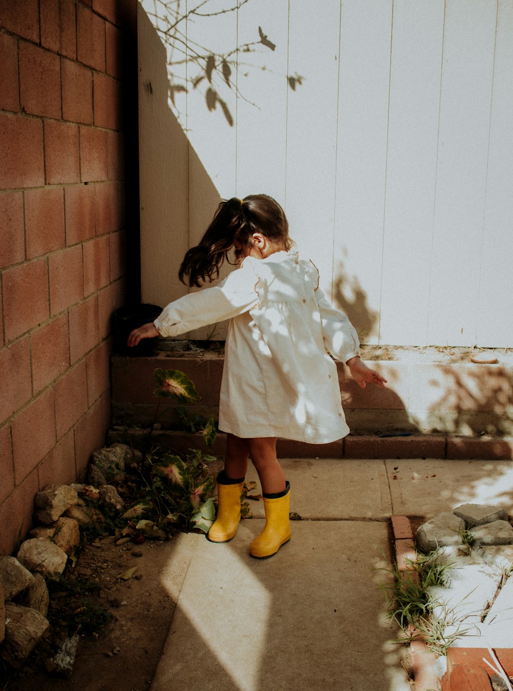 menina no casaco branco e botas amarelas de pé ao lado da parede de tijolo marrom
