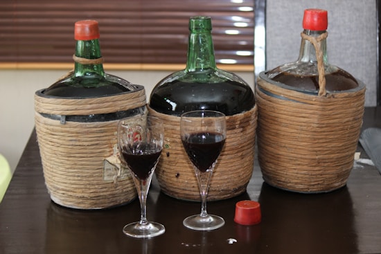 Three large wicker-wrapped bottles with green glass tops are placed on a dark wooden table, accompanied by two wine glasses filled with a dark red liquid. The bottles have a rustic appearance, with two featuring red caps. The table is set indoors with a blurred background.