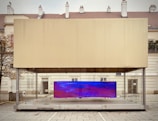 A modern art display encased in a transparent box, situated in an urban setting. The installation features a large, rectangular digital screen displaying abstract blue and purple visuals. The structure is positioned on a paved plaza, with a backdrop of residential or official buildings and a leafless tree to the side.