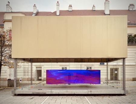 A modern art display encased in a transparent box, situated in an urban setting. The installation features a large, rectangular digital screen displaying abstract blue and purple visuals. The structure is positioned on a paved plaza, with a backdrop of residential or official buildings and a leafless tree to the side.