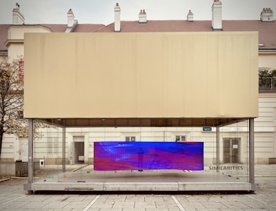 A modern art display encased in a transparent box, situated in an urban setting. The installation features a large, rectangular digital screen displaying abstract blue and purple visuals. The structure is positioned on a paved plaza, with a backdrop of residential or official buildings and a leafless tree to the side.