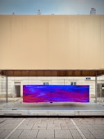 A modern urban art installation featuring a large, rectangular screen displaying an abstract digital pattern with vivid blue and red colors. The screen is mounted on a glass surface within a minimalist architectural setting, characterized by clean lines and neutral-colored walls. The scene includes a paved sidewalk in the foreground.