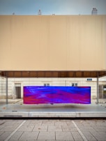 A modern urban art installation featuring a large, rectangular screen displaying an abstract digital pattern with vivid blue and red colors. The screen is mounted on a glass surface within a minimalist architectural setting, characterized by clean lines and neutral-colored walls. The scene includes a paved sidewalk in the foreground.