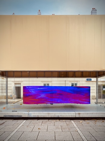 A modern urban art installation featuring a large, rectangular screen displaying an abstract digital pattern with vivid blue and red colors. The screen is mounted on a glass surface within a minimalist architectural setting, characterized by clean lines and neutral-colored walls. The scene includes a paved sidewalk in the foreground.
