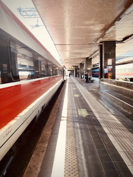 A train platform with a modern train on the left side, showing its sleek red and white design. The platform is mostly empty, with a few people visible in the distance. Overhead, there are metal beams and lights, with signs and advertisements along the walls. The lighting casts a warm, slightly muted glow.