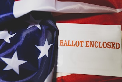 An envelope with the words 'Ballot Enclosed' printed in bold orange text lies alongside a fabric displaying part of an American flag with white stars on a blue background and red stripes.