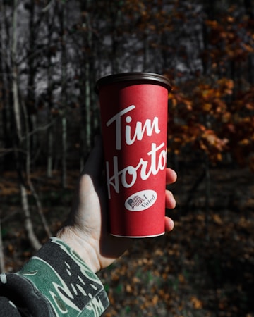 A hand wearing a camouflage jacket sleeve holds a red Tim Hortons coffee cup. The cup prominently displays the words 'Tim Hortons' and features a sticker with an American flag and the words 'I Voted'. The background consists of a forest scene with trees and a mix of greenery and fallen leaves.
