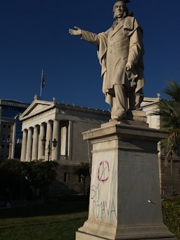 A statue of a person with one arm extended is prominently positioned in front of a classical building with columns. The pedestal of the statue has graffiti, including an anarchy symbol and the words 'Bijî Rojava'. The background features trees and a building with a flag.