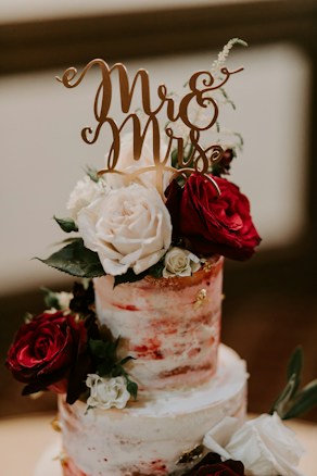 A two-tiered semi-naked wedding cake adorned with red and white roses. The cake is topped with a gold 'Mr & Mrs' topper, and decorative leaves and small white flowers are arranged around the roses. The color palette is elegant and romantic.