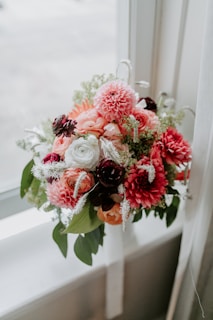 A beautifully arranged bouquet of flowers featuring a mix of pink dahlias, creamy white roses, and dark maroon blooms, accented with delicate white and green foliage. The arrangement is placed on a windowsill, allowing natural light to enhance the colors.