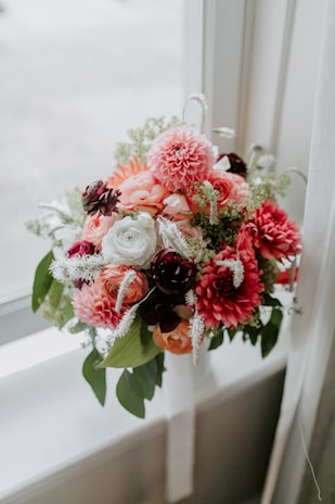 A beautifully arranged bouquet of flowers featuring a mix of pink dahlias, creamy white roses, and dark maroon blooms, accented with delicate white and green foliage. The arrangement is placed on a windowsill, allowing natural light to enhance the colors.