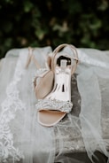 A pair of elegant, open-toed high-heeled shoes adorned with sparkling embellishments rests on a soft lace fabric. The delicate lace features intricate floral patterns, adding a sophisticated touch to the scene. The background consists of green foliage, giving a natural and serene setting.