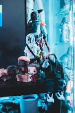 A collection of various action figures and toys are arranged on shelves, with a prominent figure resembling a bounty hunter in armor. Additional figures include stylized dolls with exaggerated features like large heads. The scene includes bright colors and a glowing blue ambient light.