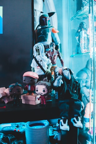 A collection of various action figures and toys are arranged on shelves, with a prominent figure resembling a bounty hunter in armor. Additional figures include stylized dolls with exaggerated features like large heads. The scene includes bright colors and a glowing blue ambient light.