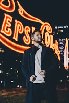 A person stands confidently in front of a brightly lit neon sign at night. The sign, featuring bold lettering and vibrant colors, illuminates the scene with a warm glow. The background is dark with city lights softly visible, adding to the urban atmosphere.