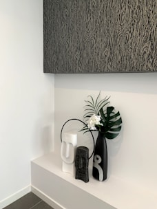 A modern minimalist decorative arrangement featuring three vases on a white shelf. The vases are of varying sizes and colors, including black, white, and gray, with abstract human-like features. A large black vase holds a green palm leaf and small white flowers. Above the arrangement is a large textured artwork in dark shades.