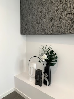 A modern minimalist decorative arrangement featuring three vases on a white shelf. The vases are of varying sizes and colors, including black, white, and gray, with abstract human-like features. A large black vase holds a green palm leaf and small white flowers. Above the arrangement is a large textured artwork in dark shades.