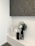 A modern minimalist decorative arrangement featuring three vases on a white shelf. The vases are of varying sizes and colors, including black, white, and gray, with abstract human-like features. A large black vase holds a green palm leaf and small white flowers. Above the arrangement is a large textured artwork in dark shades.