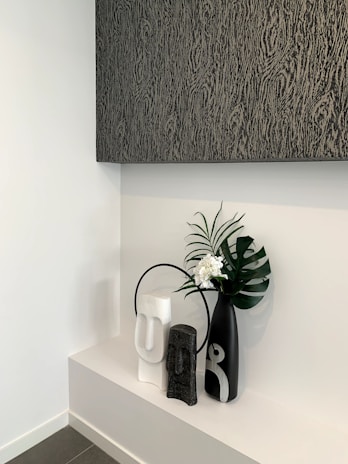 A modern minimalist decorative arrangement featuring three vases on a white shelf. The vases are of varying sizes and colors, including black, white, and gray, with abstract human-like features. A large black vase holds a green palm leaf and small white flowers. Above the arrangement is a large textured artwork in dark shades.