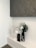 A modern minimalist decorative arrangement featuring three vases on a white shelf. The vases are of varying sizes and colors, including black, white, and gray, with abstract human-like features. A large black vase holds a green palm leaf and small white flowers. Above the arrangement is a large textured artwork in dark shades.