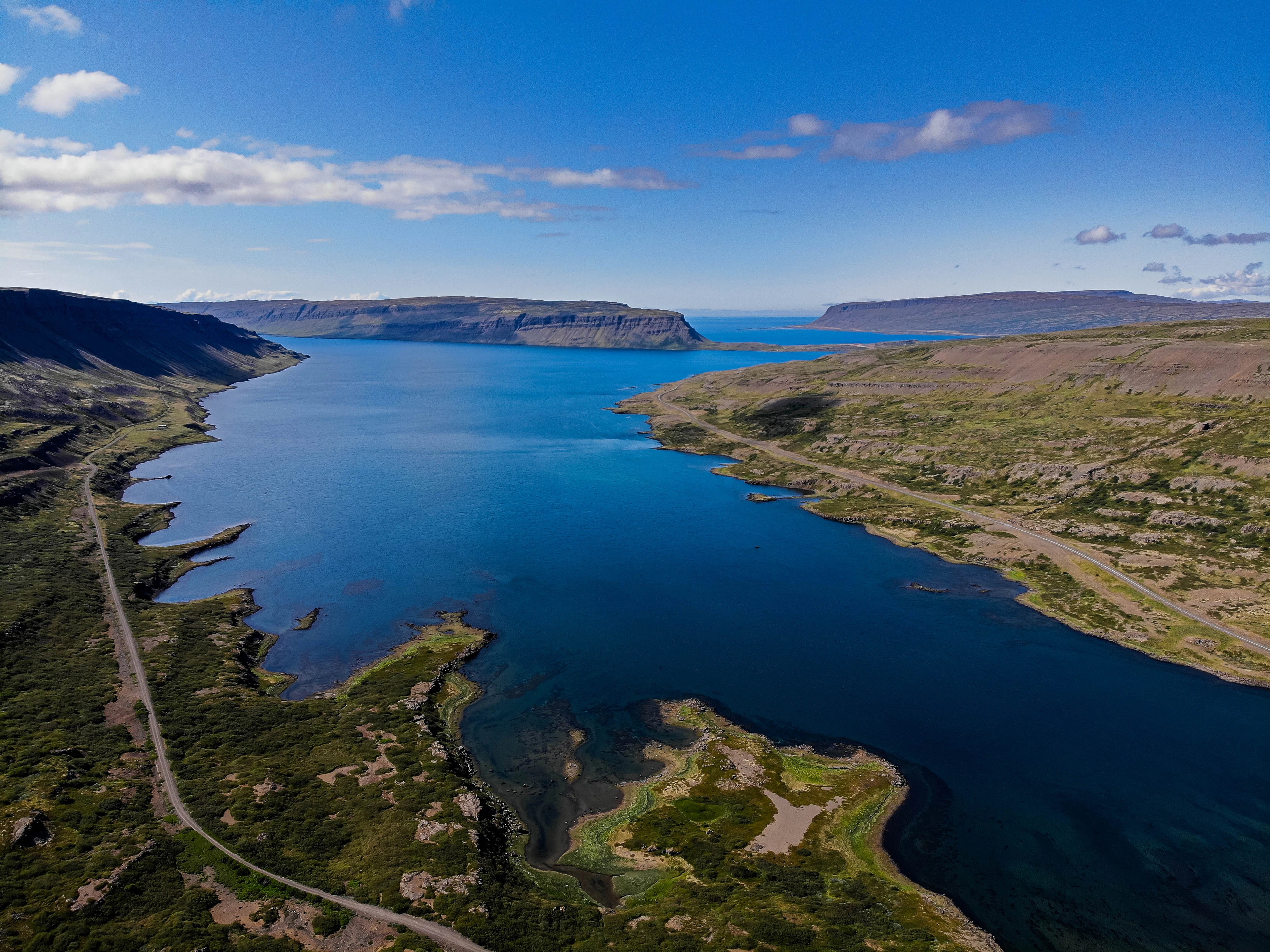 Aerial view of a long blue fjord, green shores and a road winding along the water in the Westfjords