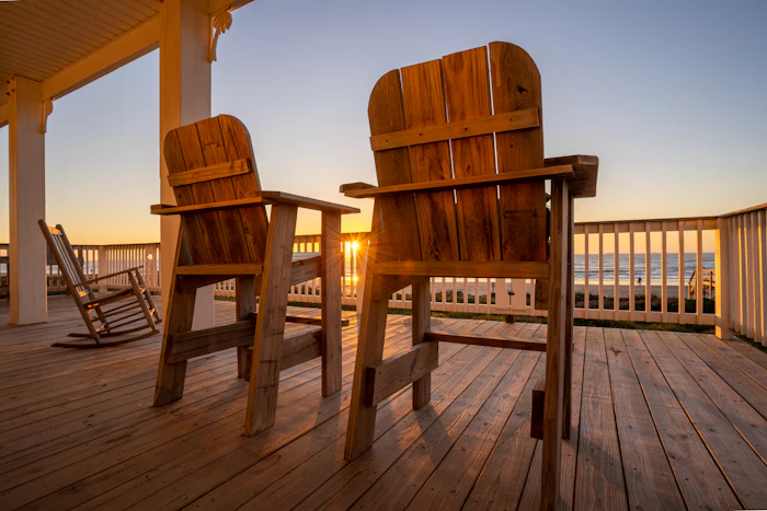Two wooden Adirondack chairs on a deck at sunset
