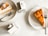 Slices of pie are served on white plates, accompanied by metal forks. Nearby, cartons labeled 'Boxed Water Is Better' are placed on a marble table surface.