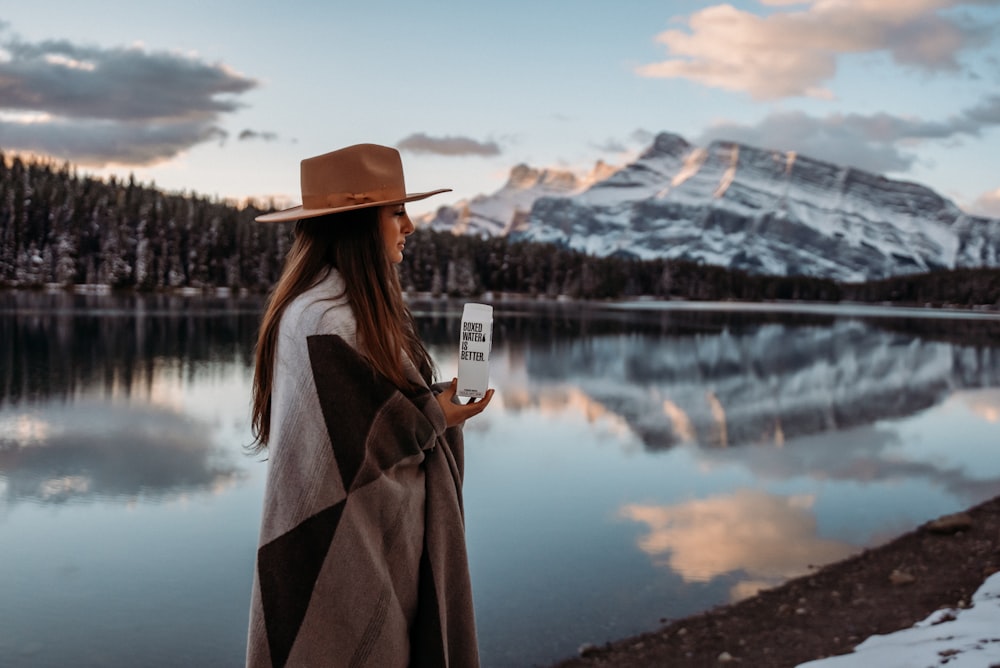 mulher no casaco marrom que veste o chapéu marrom em pé perto do lago durante o dia